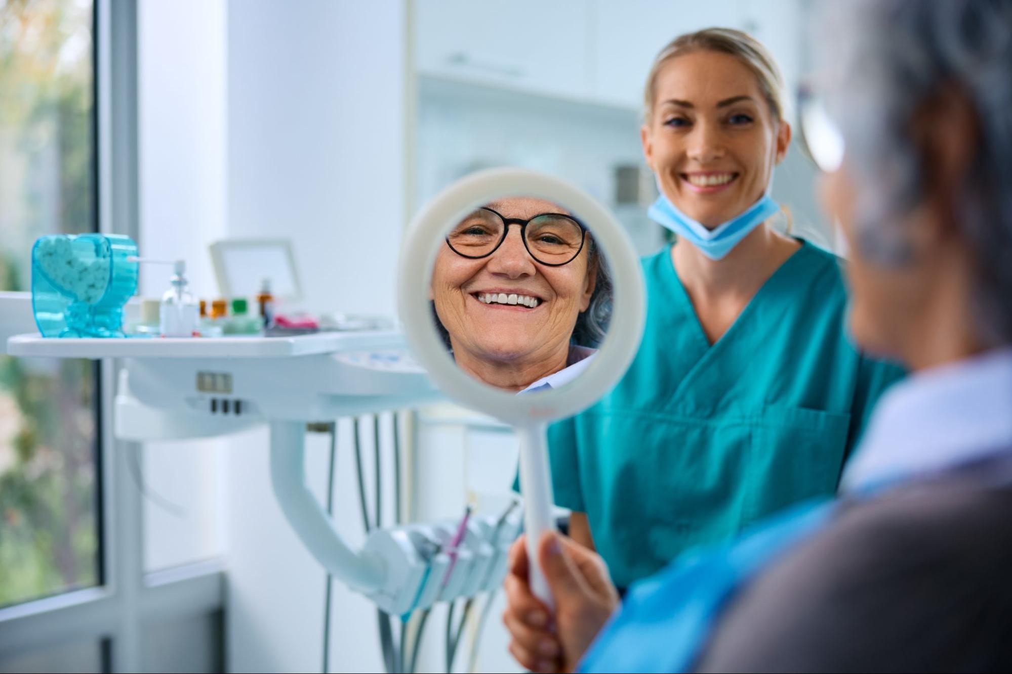 Smiling senior woman admiring her newly fitted dentures in a mirror, with her dentist standing nearby, offering guidance and support.