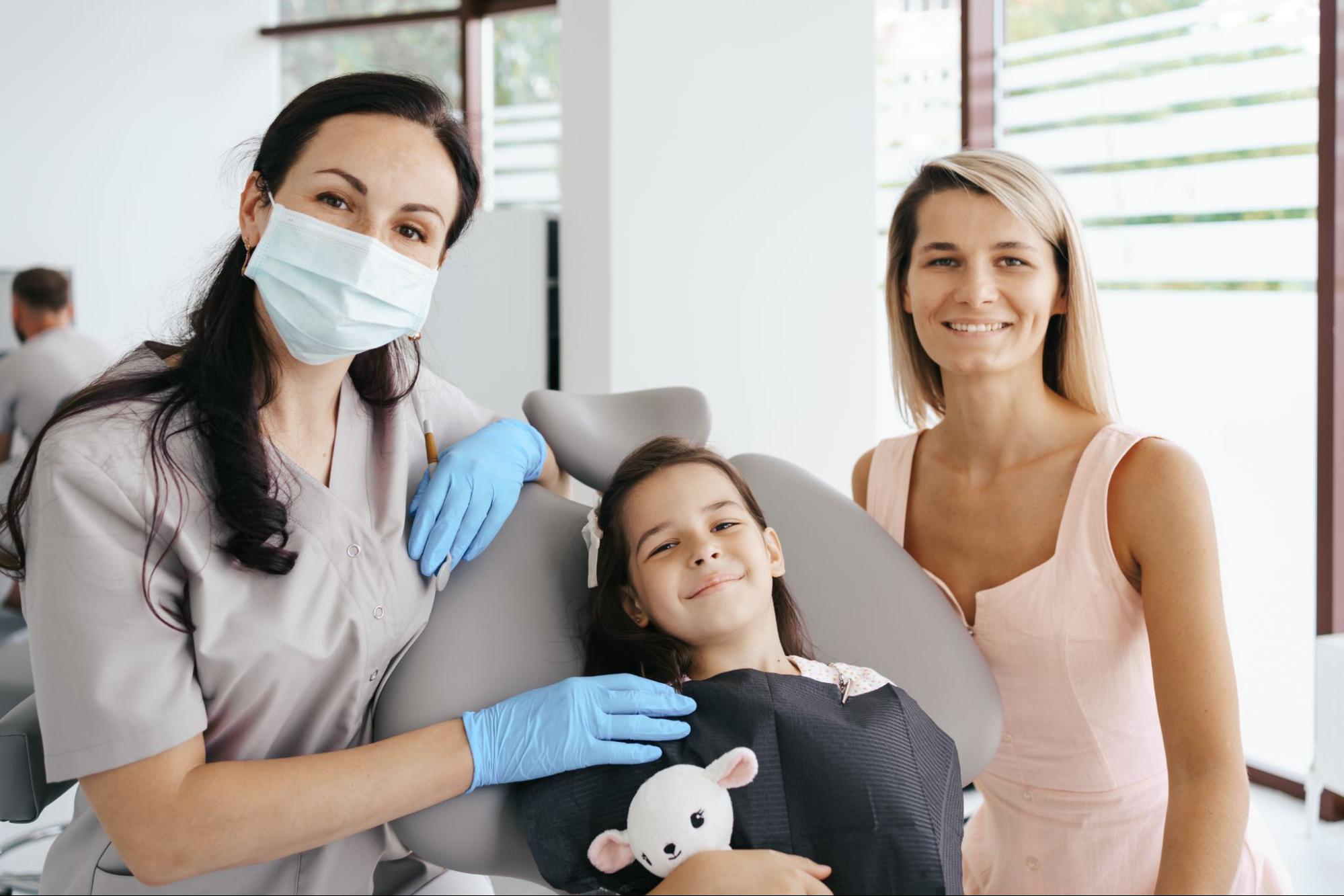 A friendly dentist poses with a young girl holding a stuffed toy and her smiling mother in a brightly lit dental clinic, creating a warm and reassuring atmosphere.
