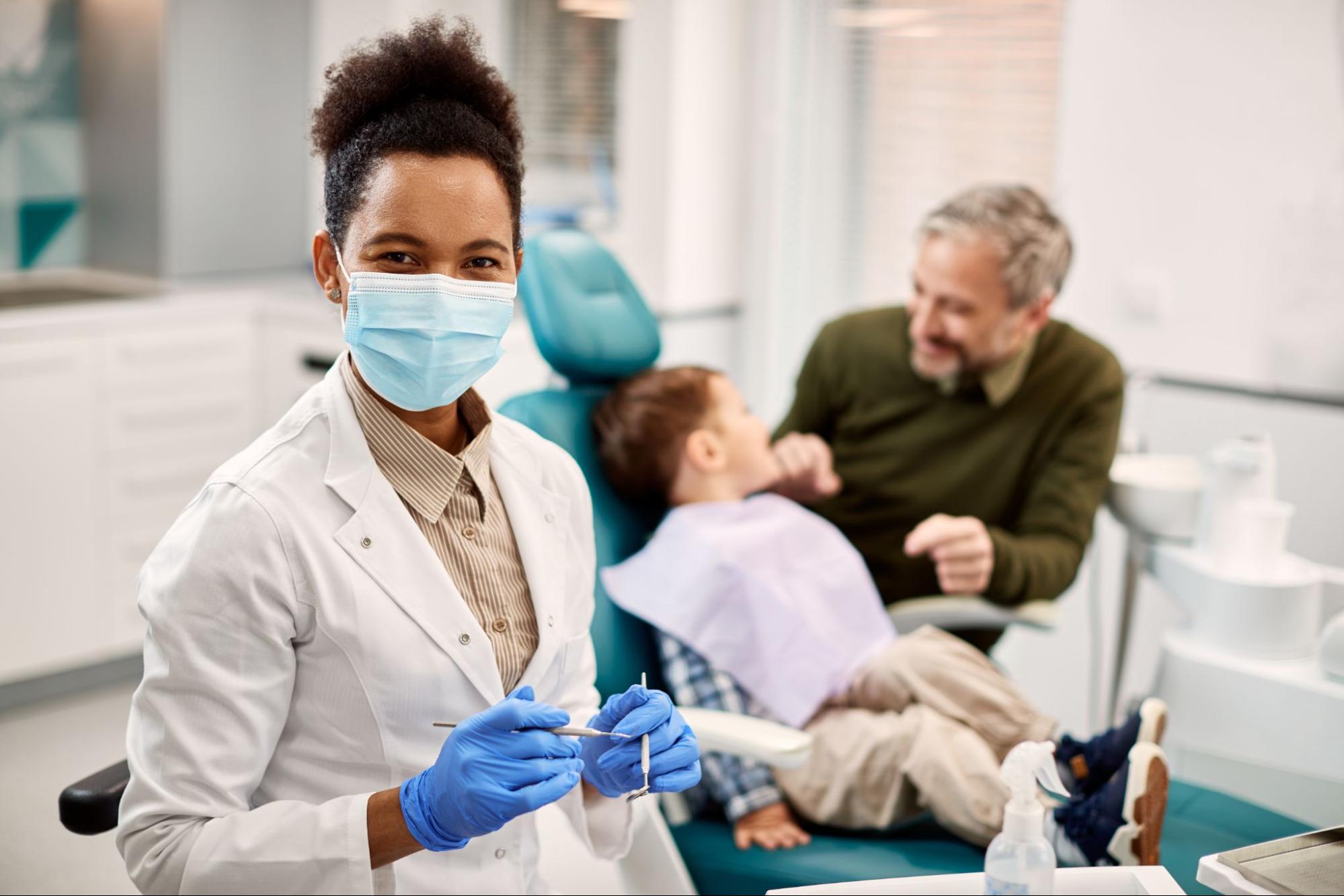 In the dental office, a friendly dentist wearing a mask and gloves smiles confidently at the camera while preparing tools while a young child sits in a dental chair with his father in the background.