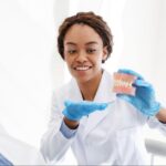 A female dentist explains the dental bridge procedure to a male patient using a plastic dental model.