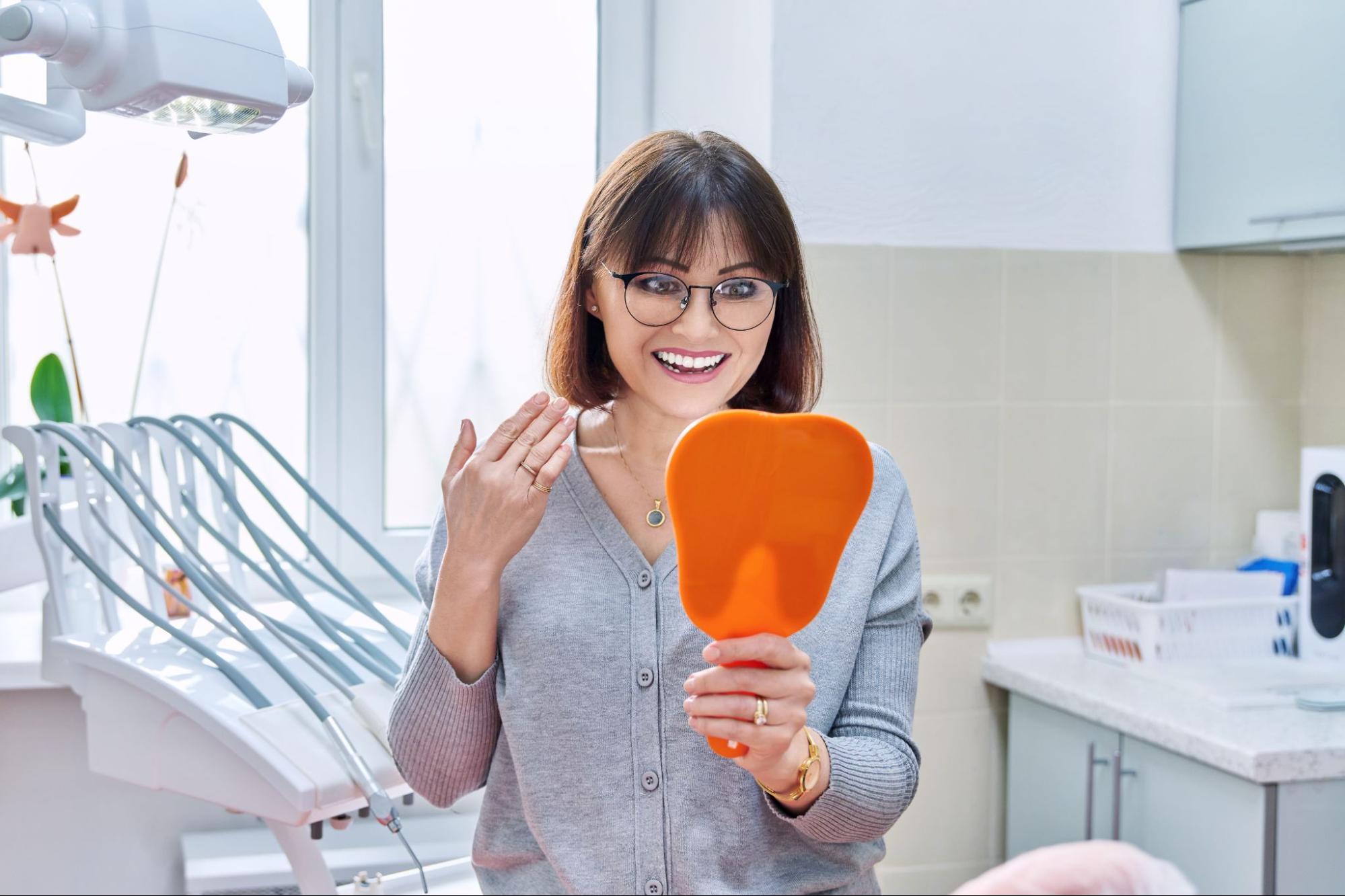 A visibly pleased female dental patient examines her dental bridges in a handheld mirror.