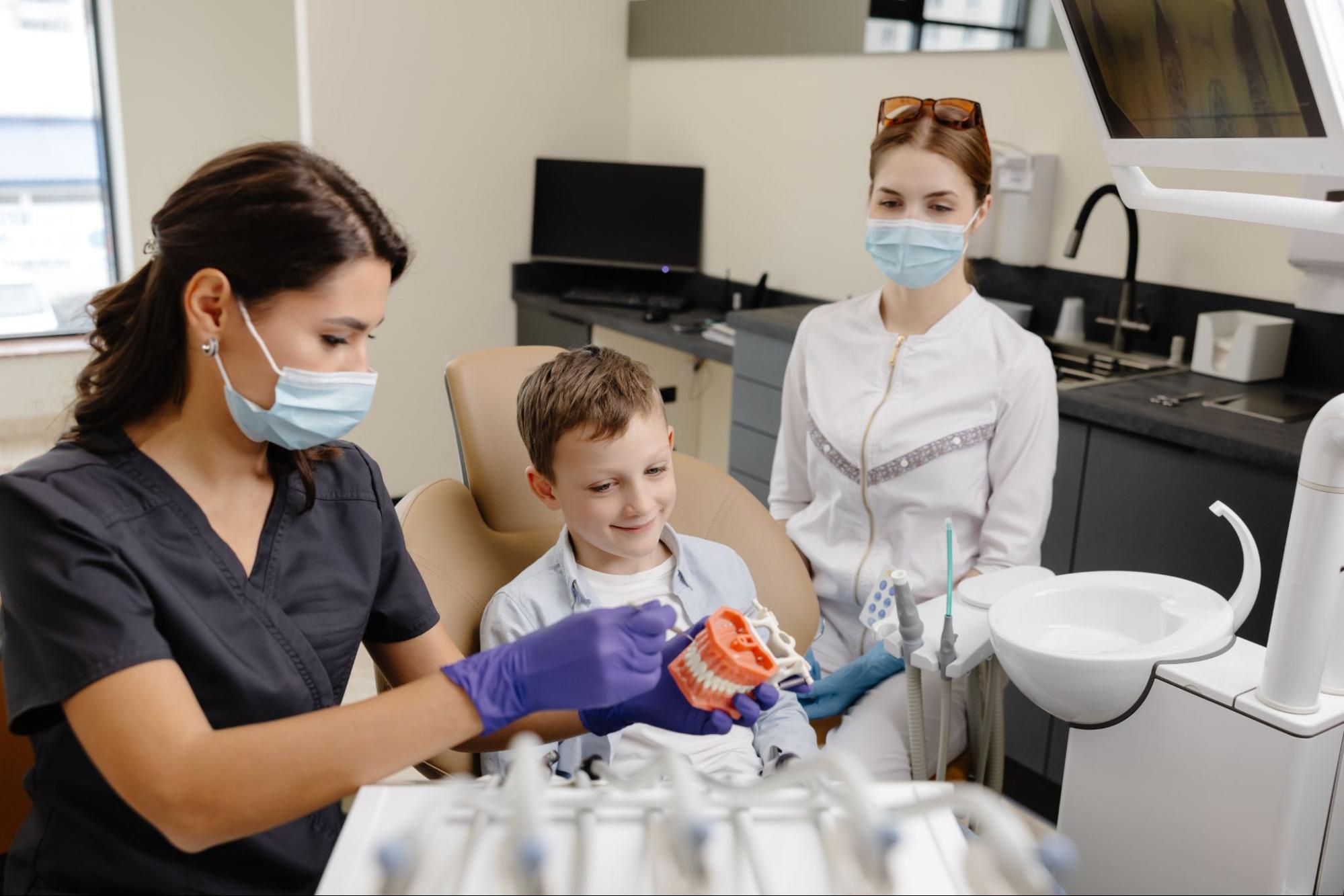 A pediatric dentist demonstrates a dental procedure using a model to a young boy while the dental assistant observes.