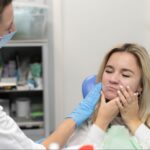 A woman holding her cheek in pain is examined by a dentist providing emergency dental care.
