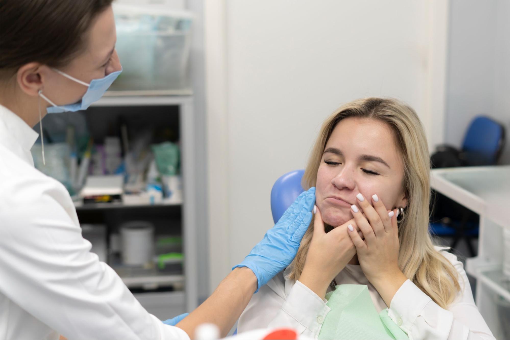 A woman holding her cheek in pain is examined by a dentist providing emergency dental care.
