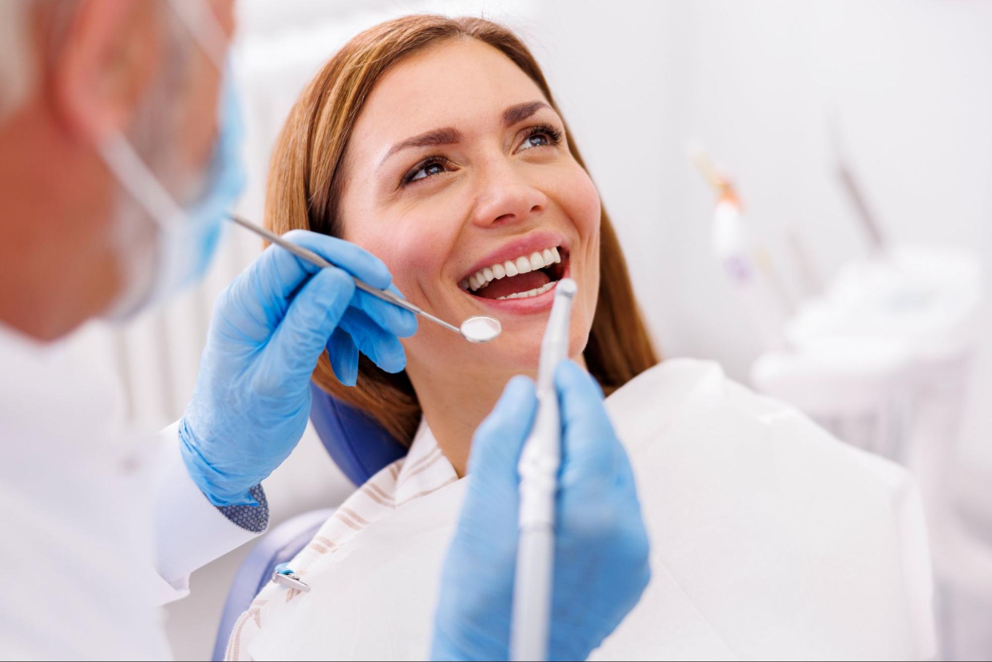 A woman smiling in an exam chair next to a dentist holding tools. 