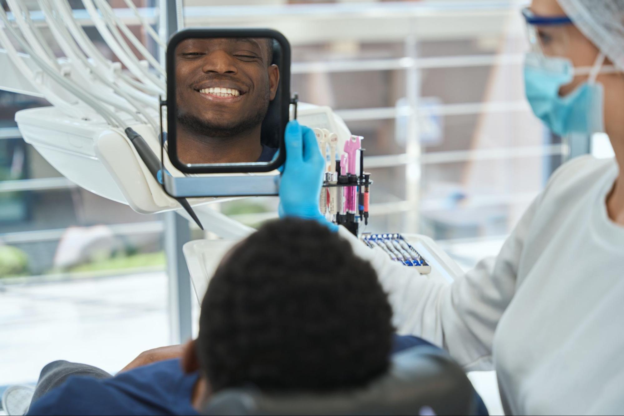 A male patient is looking at his smile in a mirror held by the dentist after teeth cleaning.