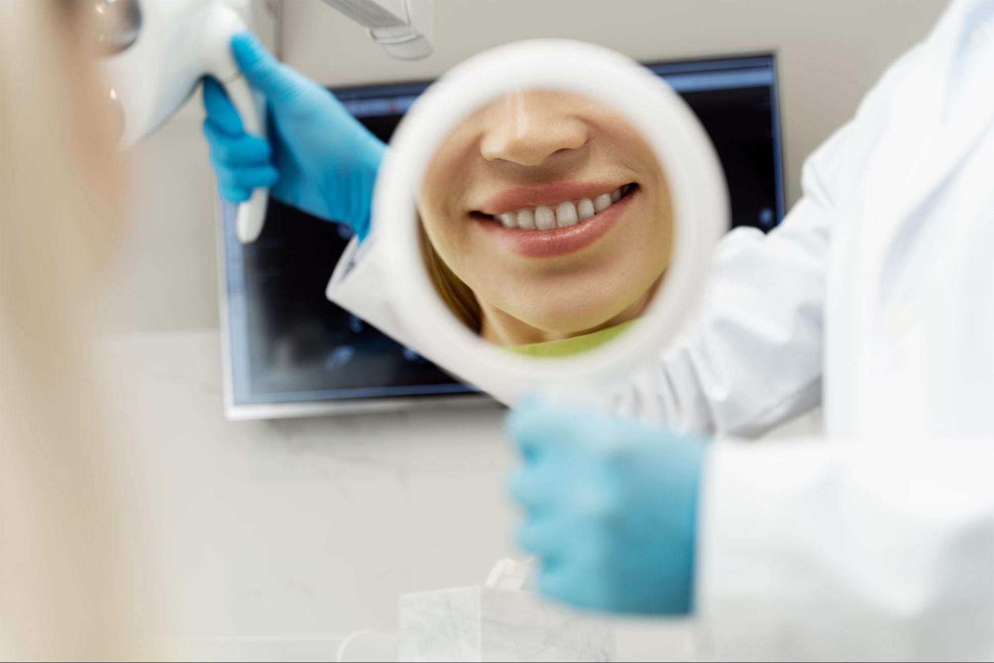 A female patient with a bright smile happily examines her teeth in a mirror after a professional teeth cleaning.