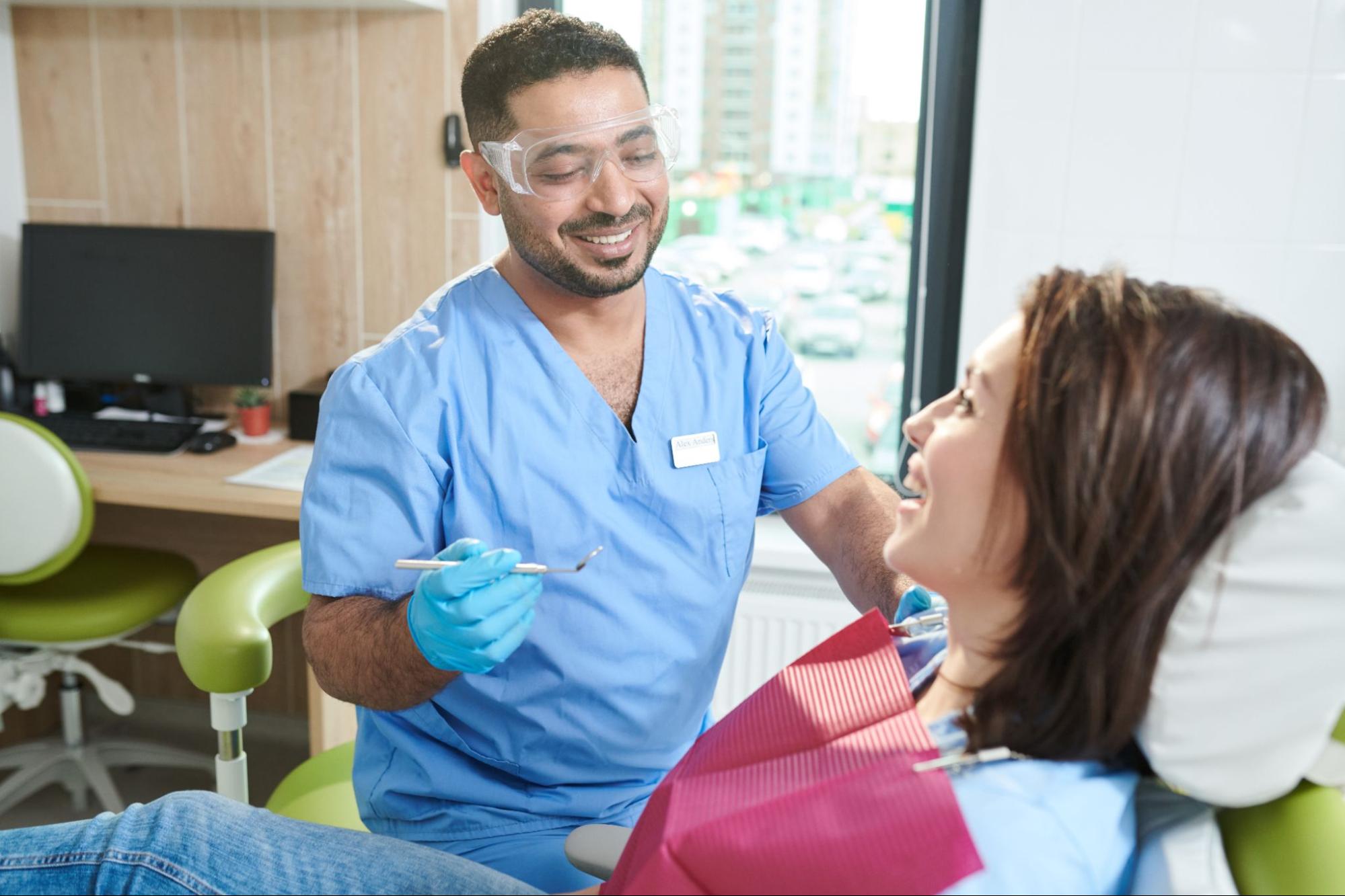 Woman in an exam chair smiling next to a male dentist in googles. 