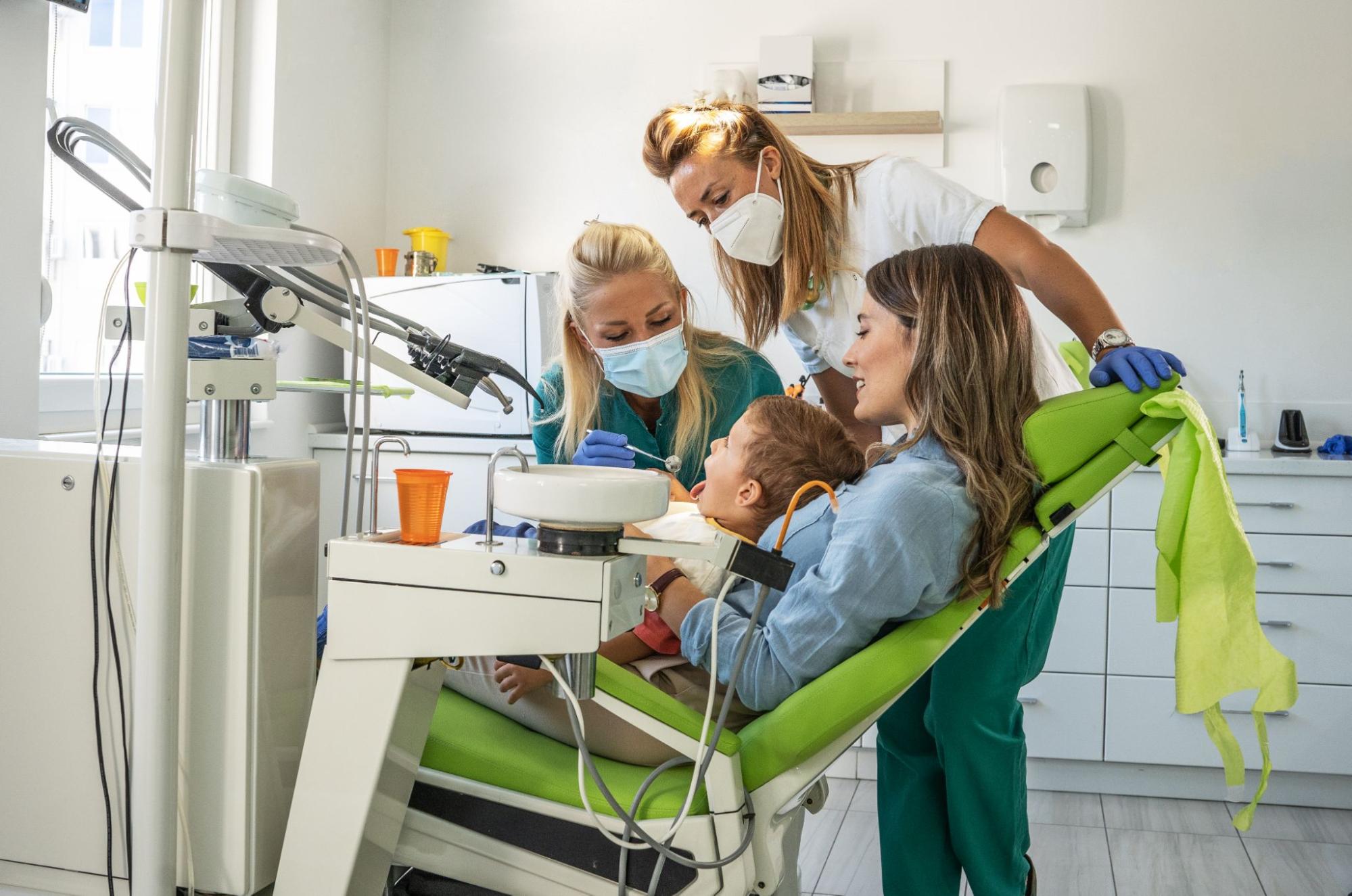 A team of dental professionals attends to a young child in a brightly lit clinic. The mother sits beside the child, ensuring a supportive and comforting environment during the procedure.