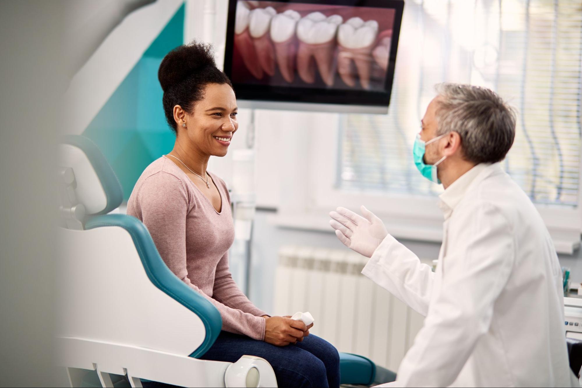 A happy Black woman discusses her dental bridge procedure with her orthodontist while viewing a 3D teeth scan on the monitor.