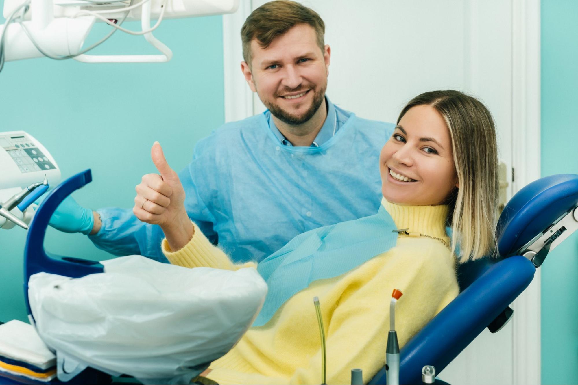A beautiful lady sitting on the dentist's chair smiles and gives a thumbs-up after successful gum disease treatment, showing confidence in her oral health.