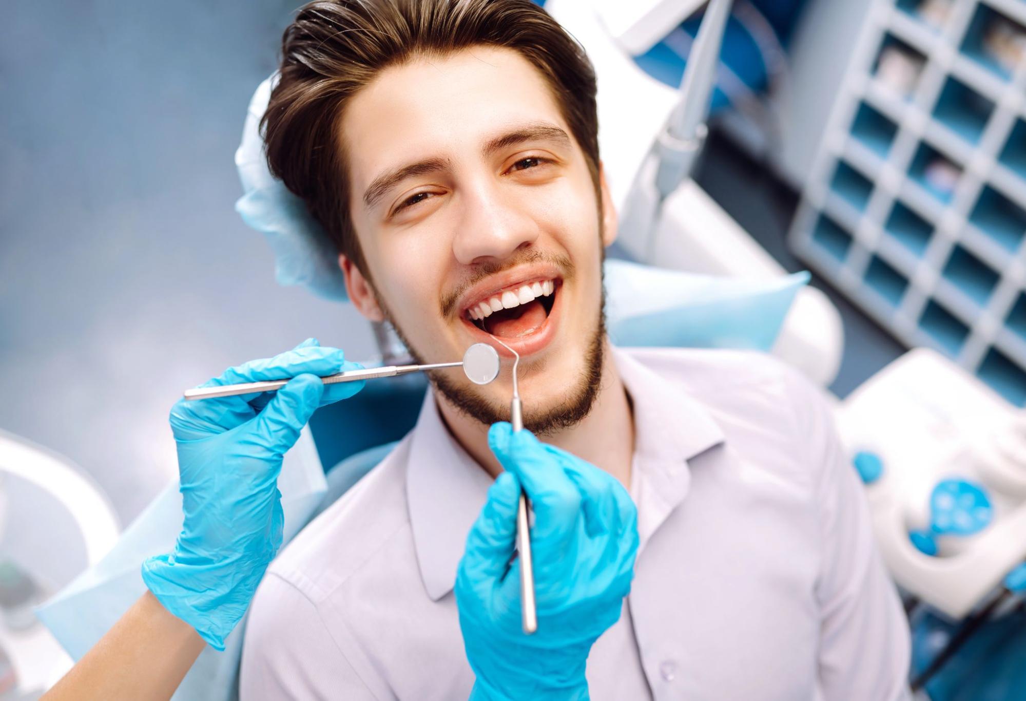 A young man in the dentist's chair undergoing a deep teeth cleaning smiles brightly as unrecognized hands hold dental tools near his mouth.
