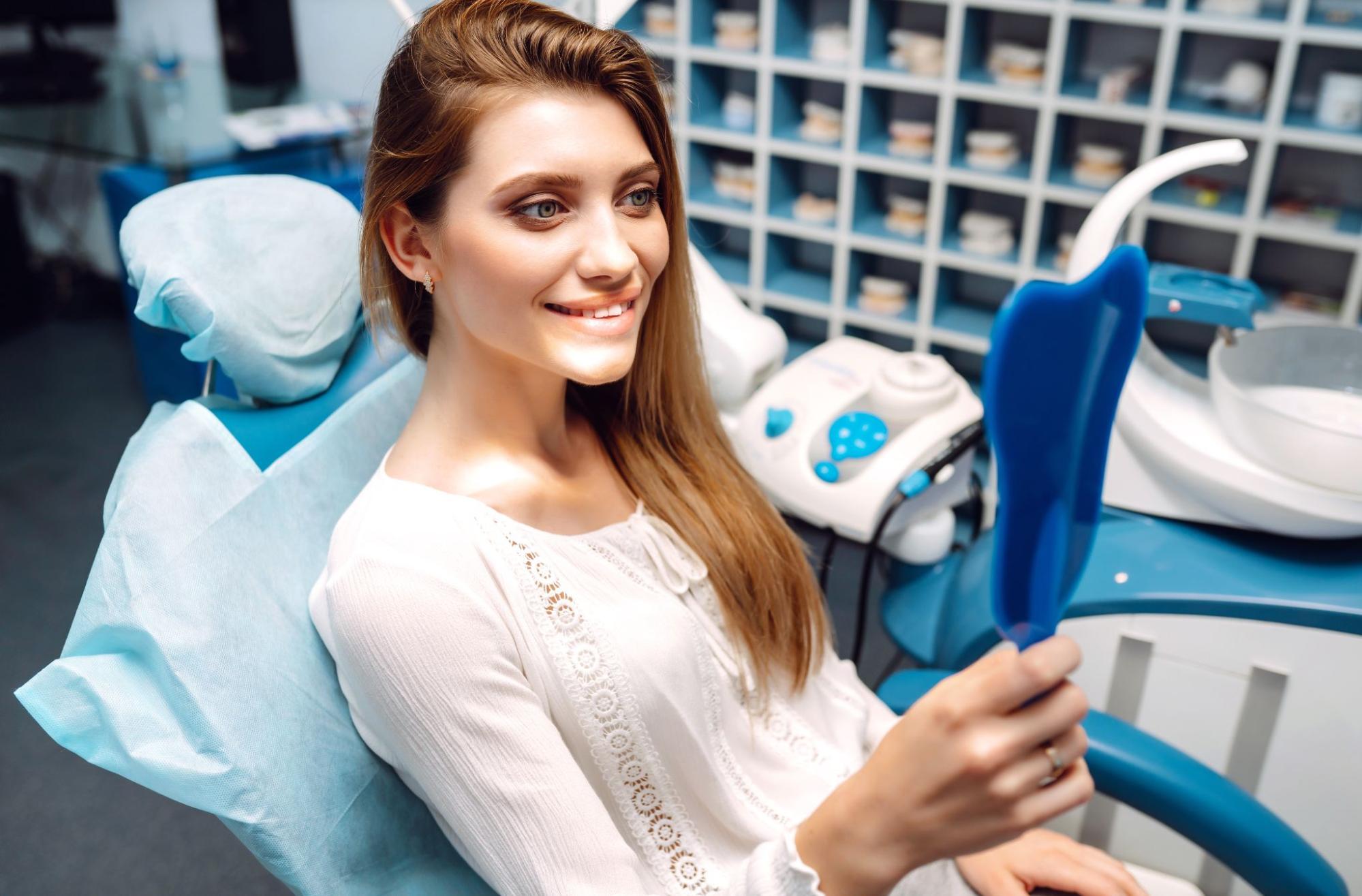 A young woman in the dentist's chair smiles and admires her brighter teeth in a mirror after a professional teeth whitening procedure.