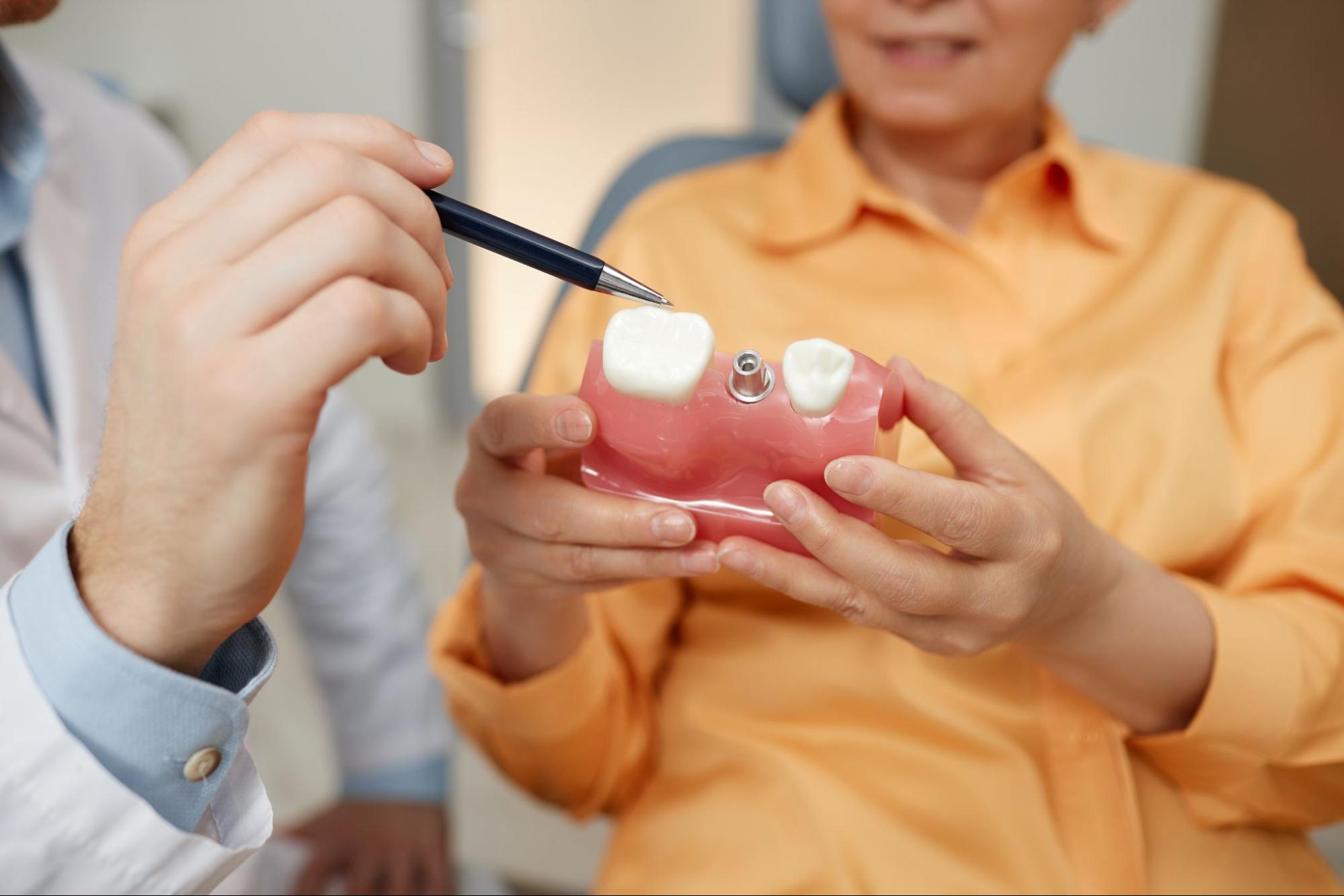 A close-up of an unrecognizable dentist pointing at a dental implant model while the patient holds it.