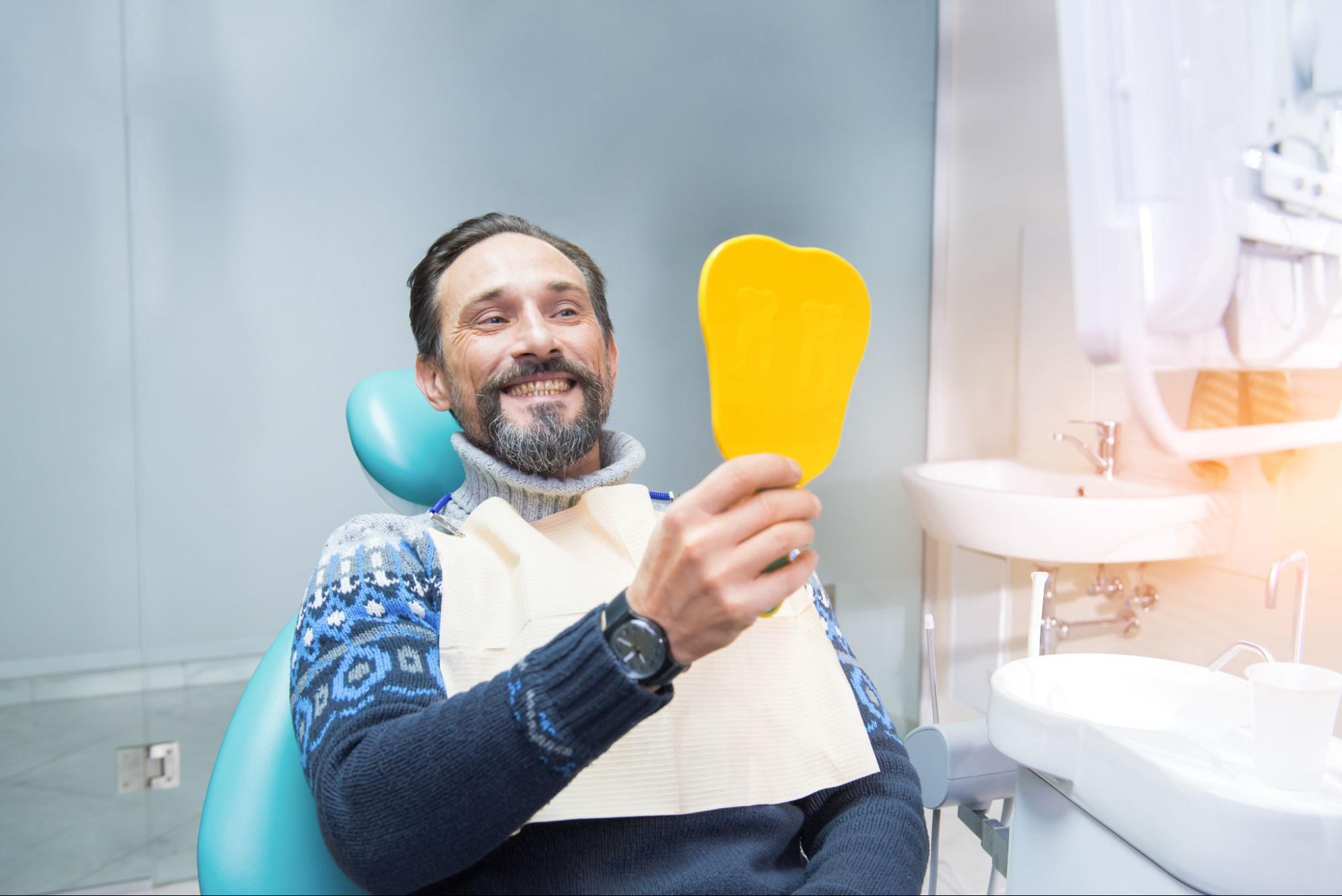  A satisfied patient in a dental chair admires his new dental bridges in a yellow mirror. 