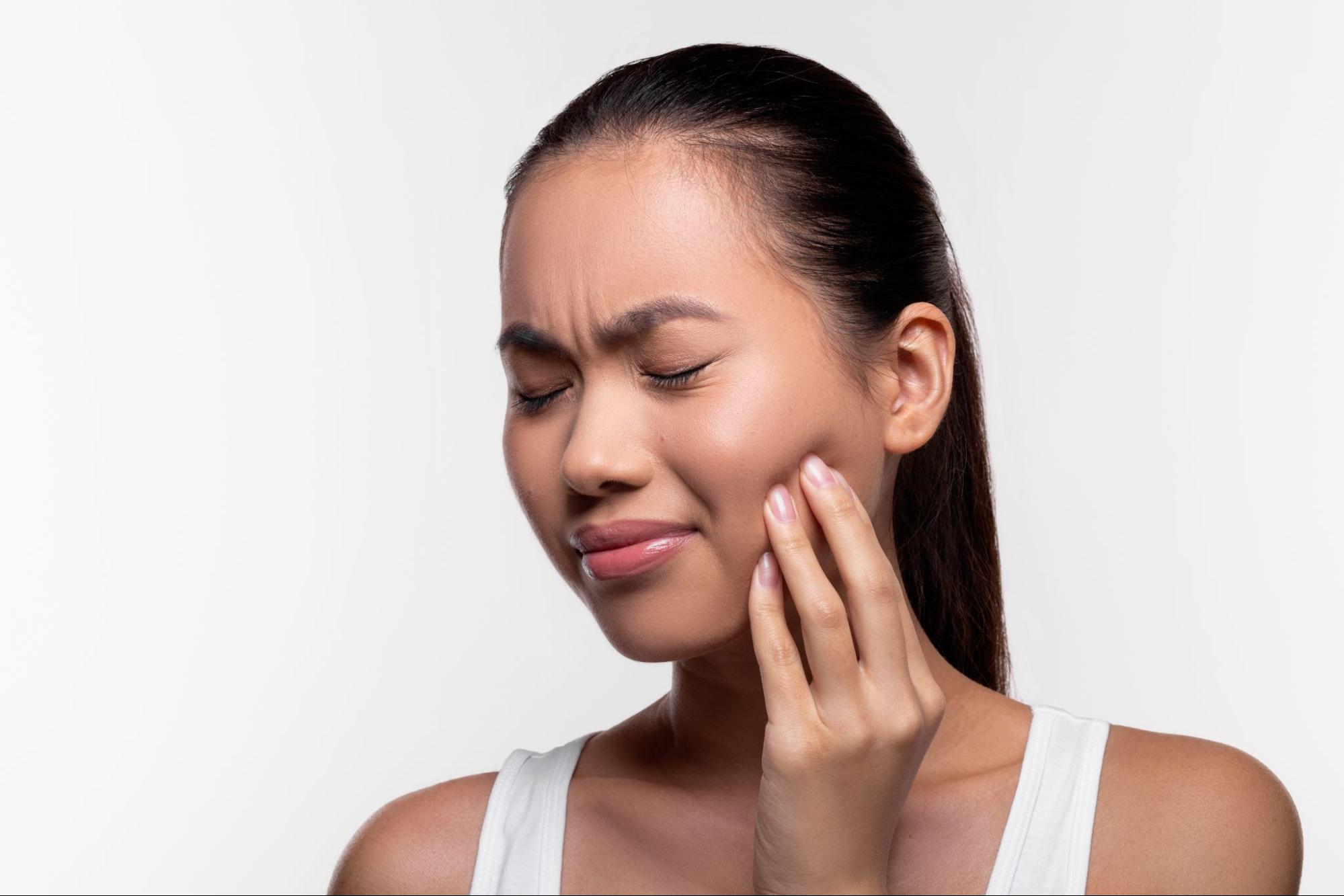 A Korean woman holds her cheek in discomfort, showing signs of TMJ disorder