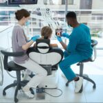 A Black male dentist presents the teeth model to the patient as the assistant sets up.