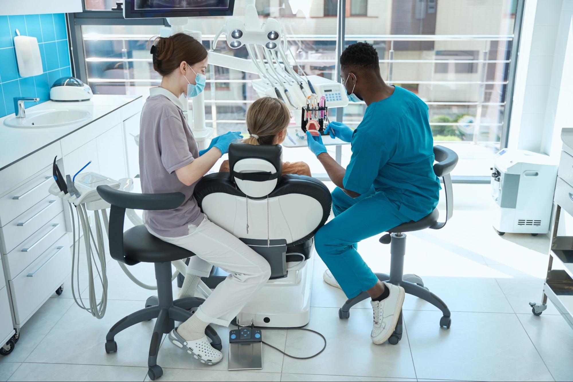 A Black male dentist presents the teeth model to the patient as the assistant sets up.