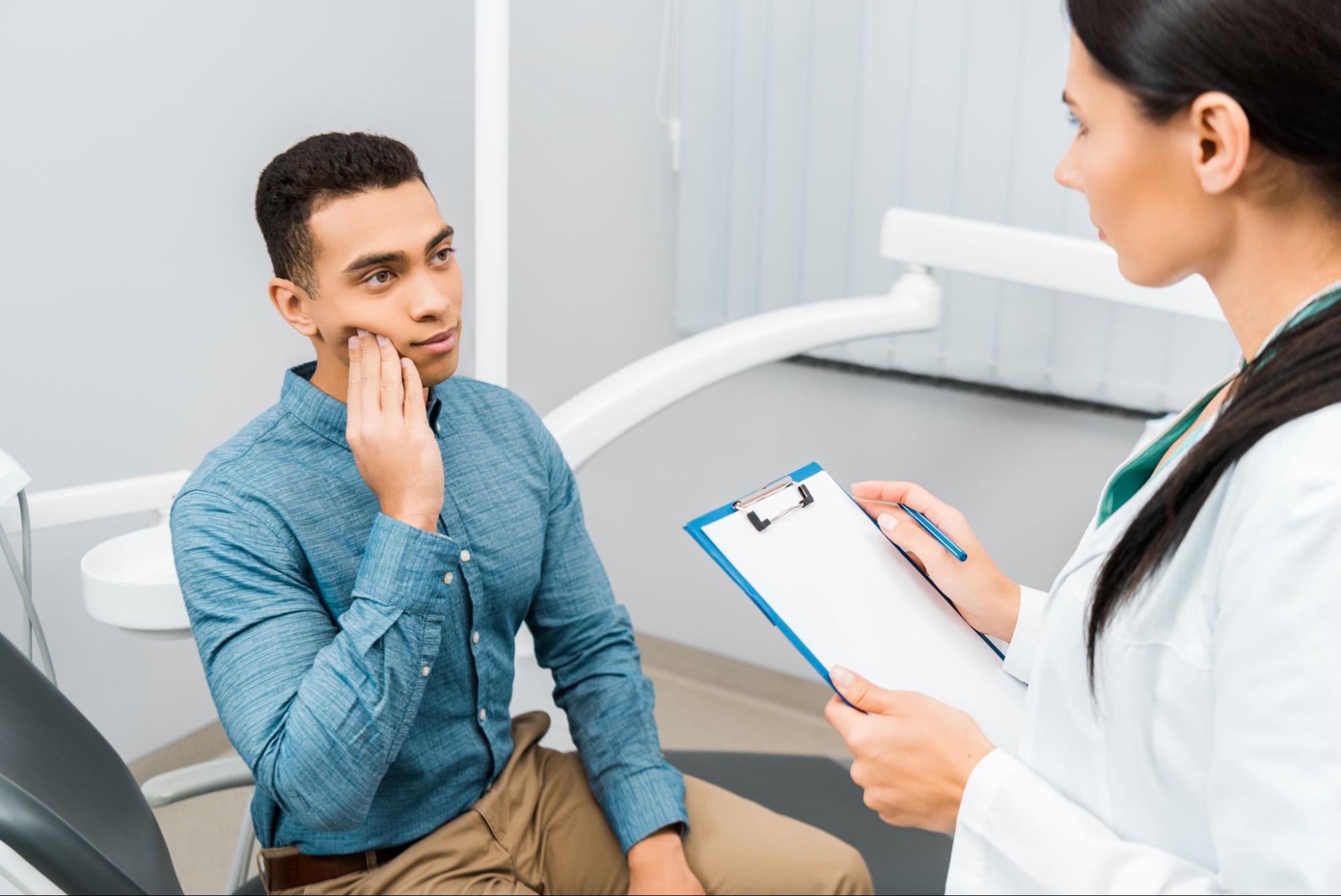 A female dentist holding a clipboard interviews a male patient with a toothache.