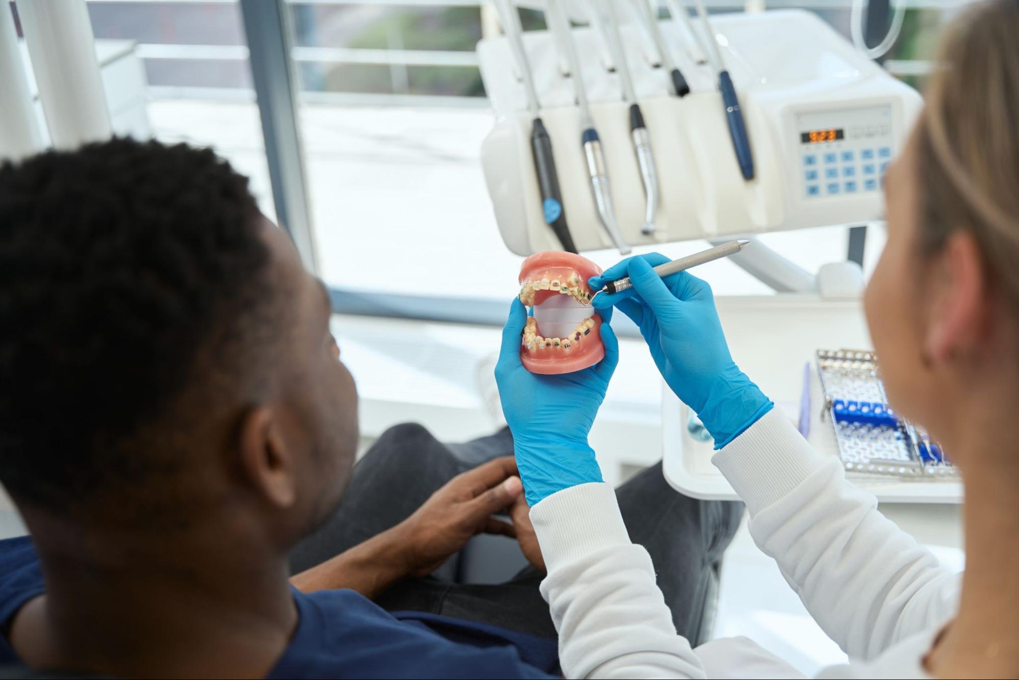 A female orthodontist uses a jaw model to explain the male patient’s dental issues.