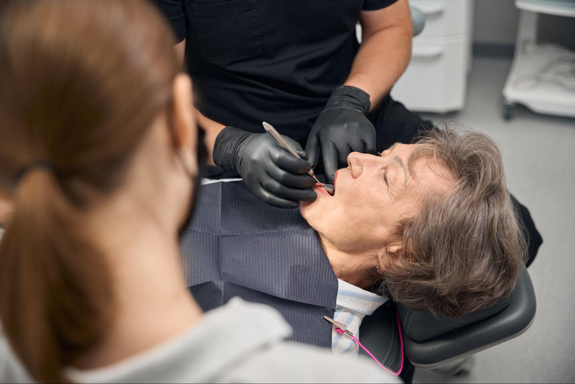 A dentist examines an elderly woman’s teeth while an assistant observes.