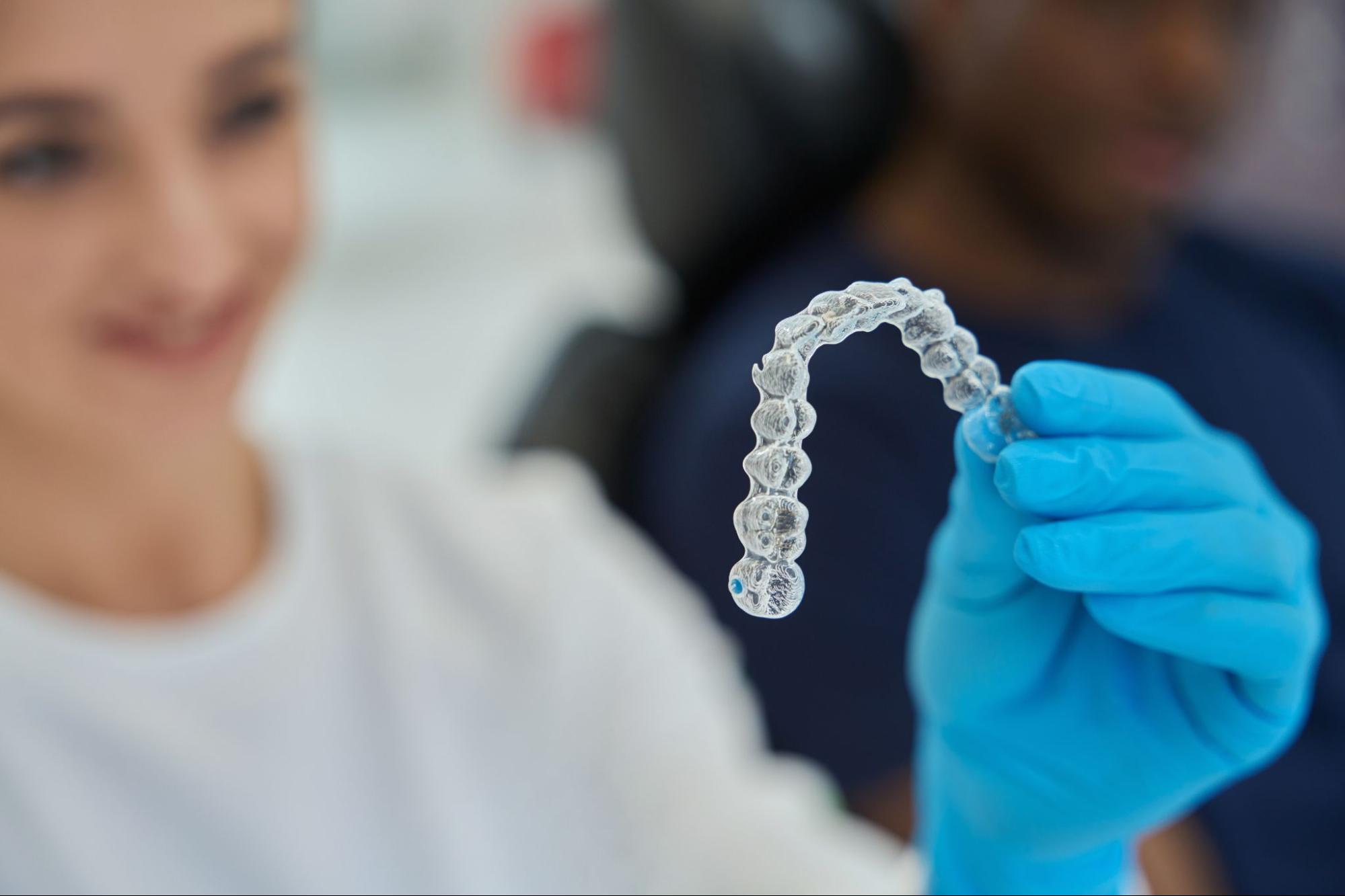 A dental technician in blue latex gloves holds a clear Invisalign® aligner.