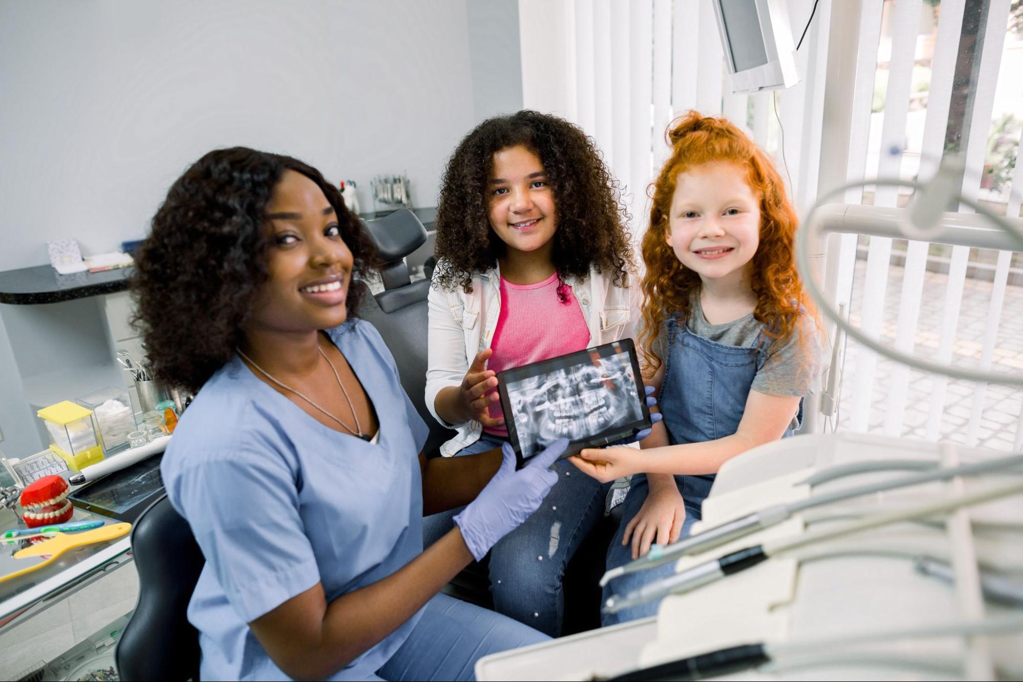 A female African pediatric dentist shows two young patients a digital dental X-ray on a tablet.