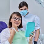 A female patient examines new dentures in the mirror while the dentist stands behind her.