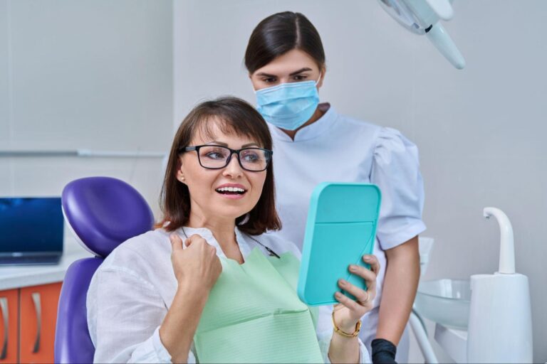 A female patient examines new dentures in the mirror while the dentist stands behind her.